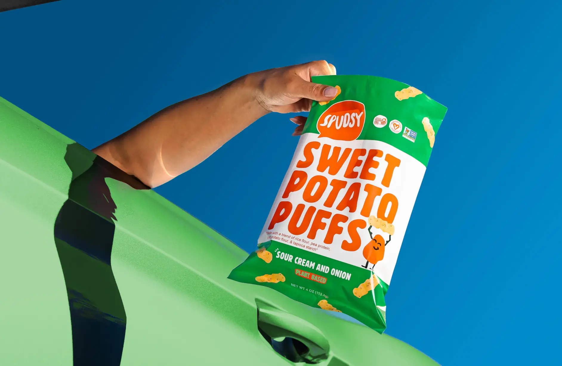 A packaging design of Spudsy potato puffs, showing a package of sudsy puffs hanging from the hand of a person in a green car, with a beautiful green color Puffs package with mix of white, orange, and yellow.